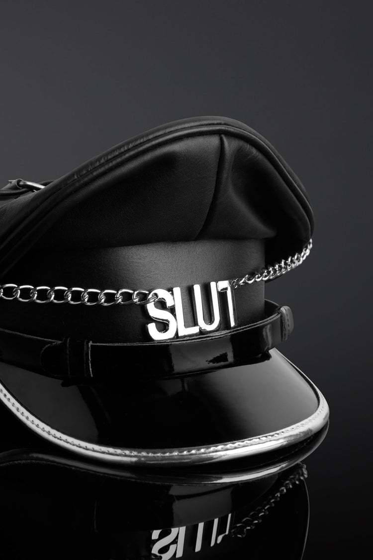 Black police hat with 'SLUT' chain on a reflective surface against a dark background