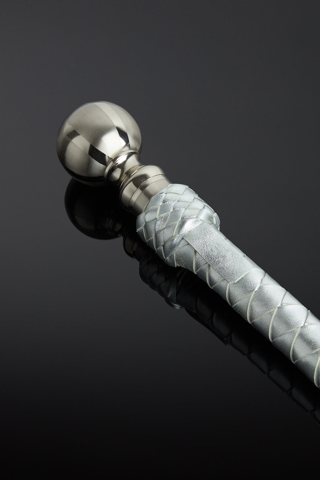 Metallic silver leather whip with a metal handle and pointed end on a reflective black surface  - closeup shot