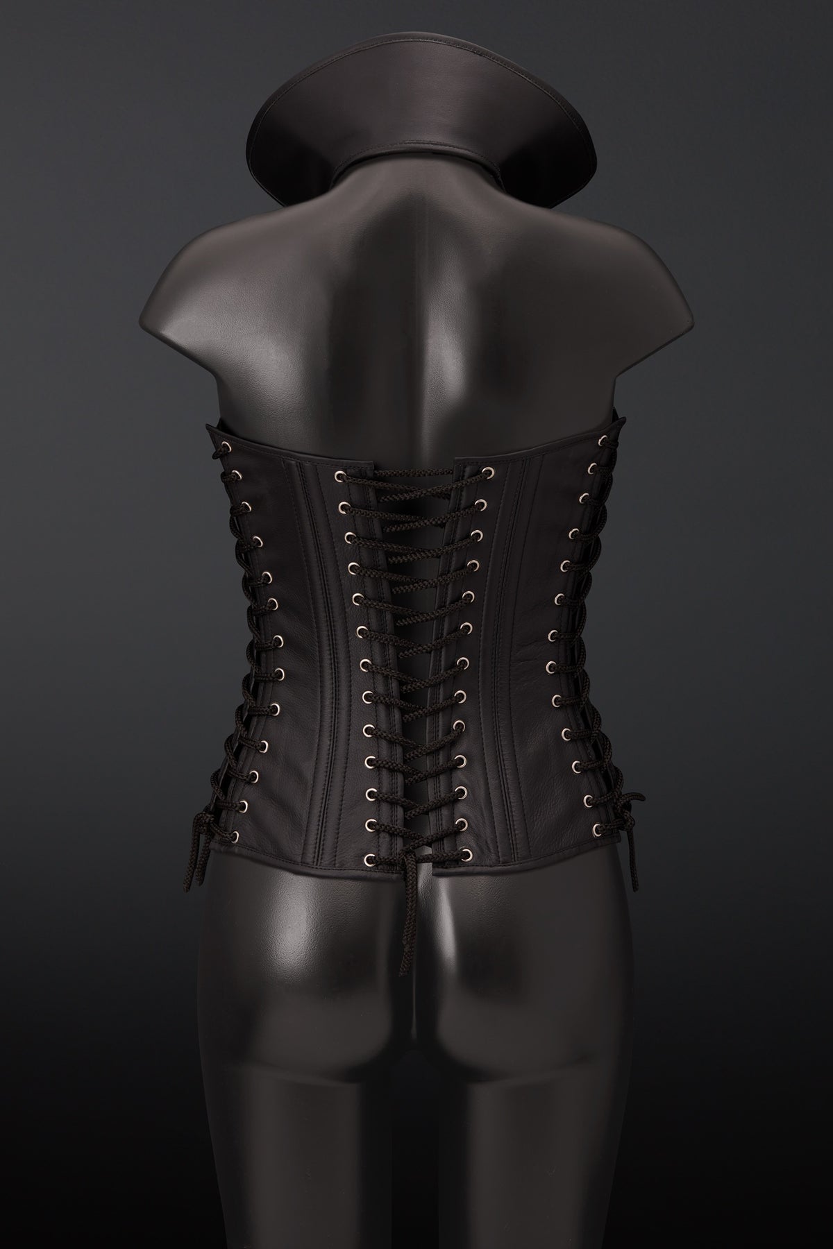 Black leather corset vest with lace-up back closure, displayed on a black mannequin from the back against a dark gray background.