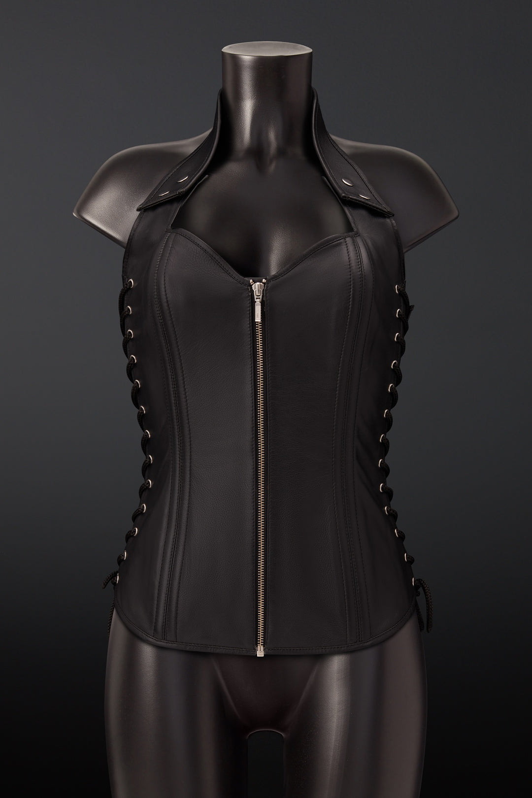 Black leather corset vest with a front zipper, shoulder straps, and side lacing, displayed on a black mannequin against a dark gray background.