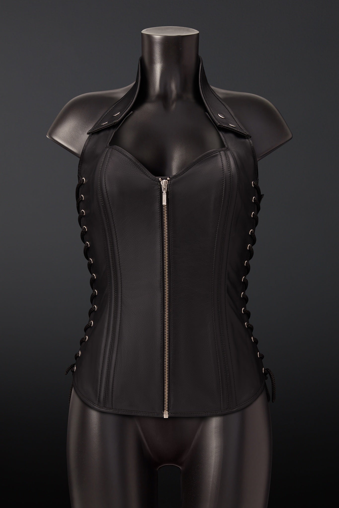 Black leather corset vest with a front zipper, shoulder straps, and side lacing, displayed on a black mannequin against a dark gray background.