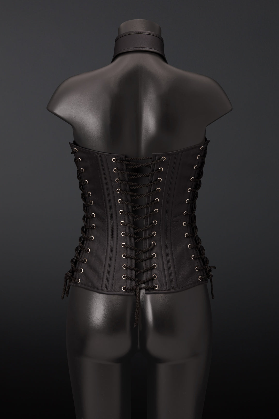 A black leather corset displayed on a black mannequin torso, viewed from the back. The corset features multiple rows of black laces threaded through brass eyelets.