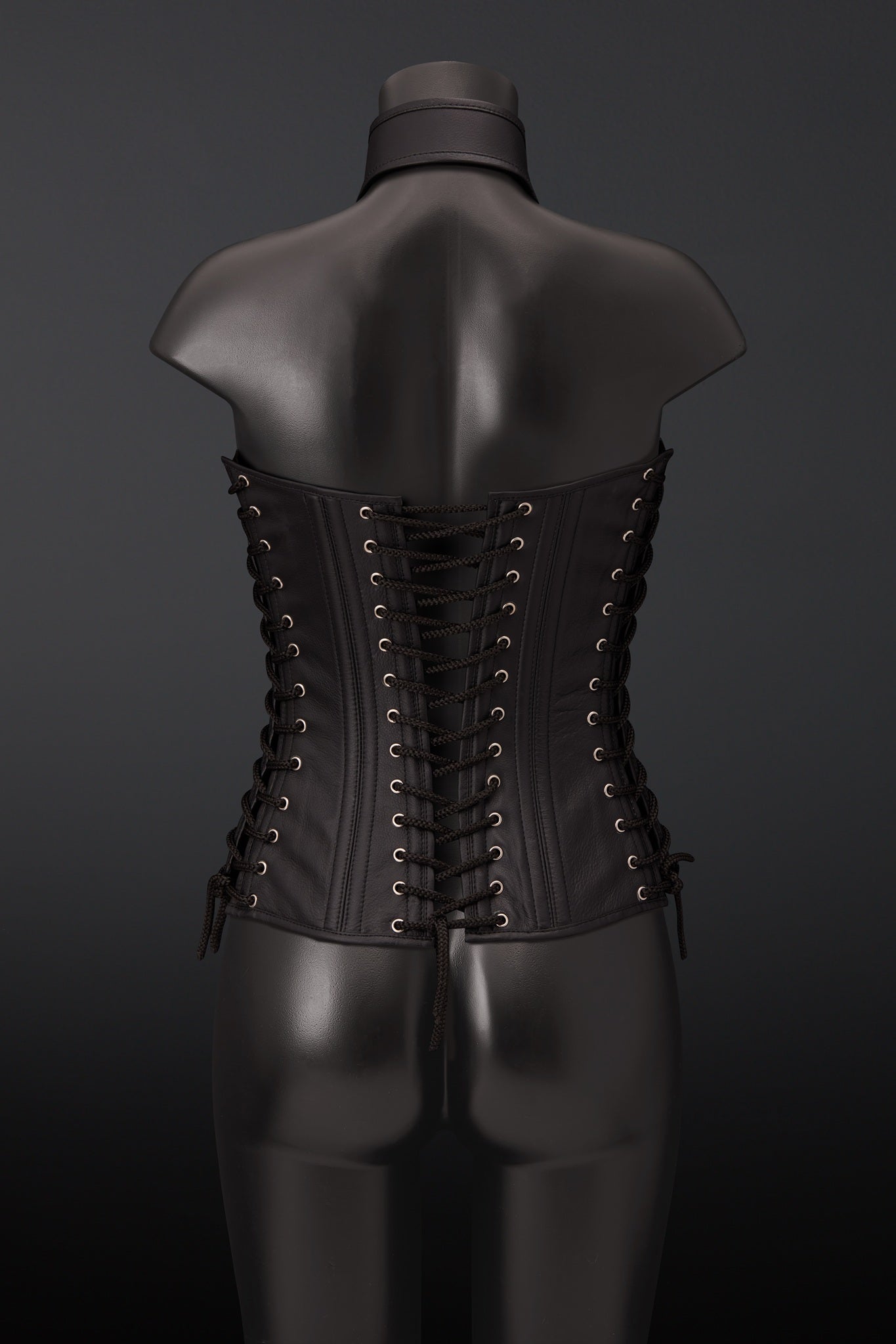 A black leather corset displayed on a black mannequin torso, viewed from the back. The corset features multiple rows of black laces threaded through brass eyelets.