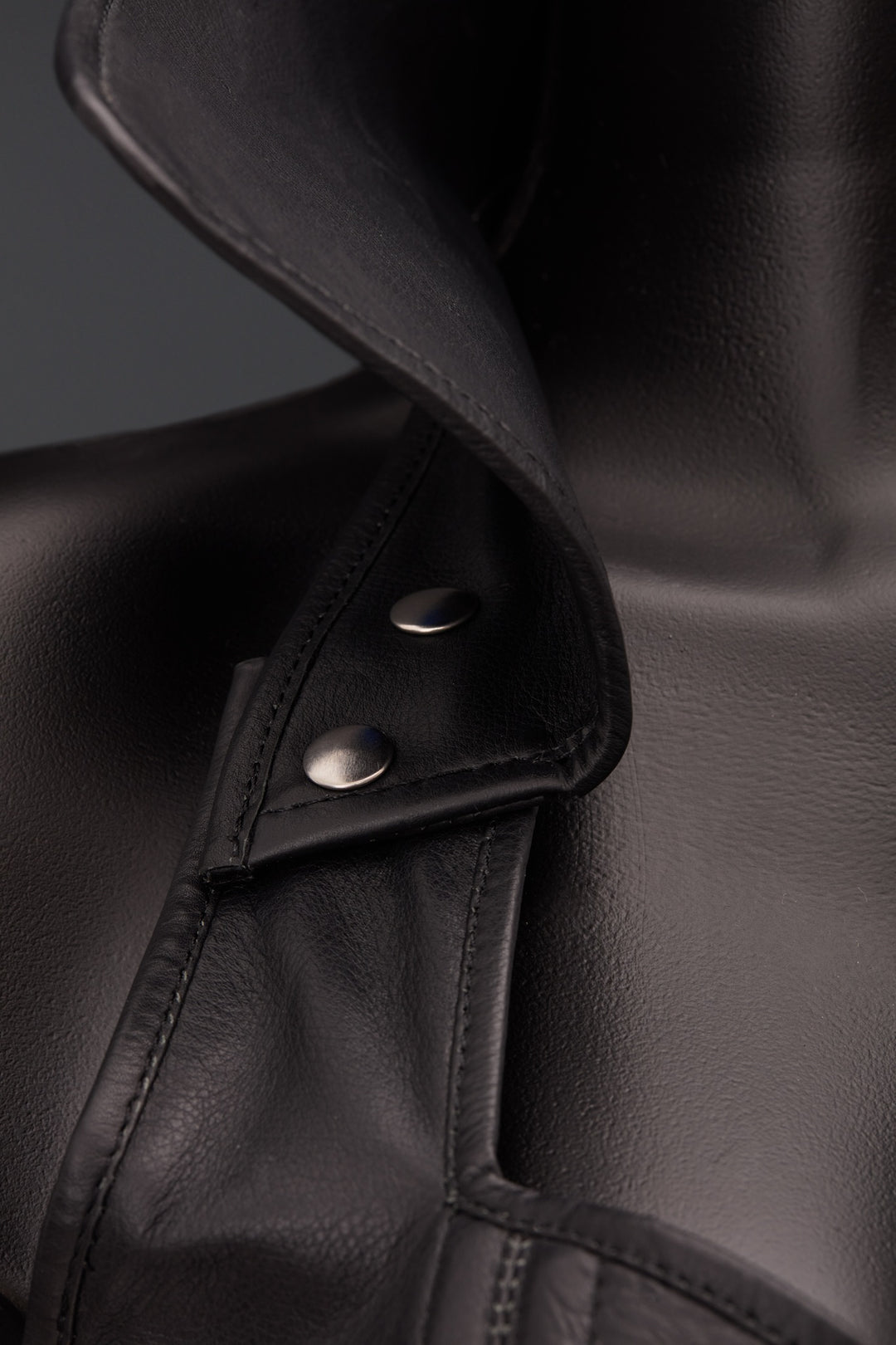 Close-up of a black leather corset with a pointed collar and silver snap button detail, shown on a dark mannequin against a dark gray background.