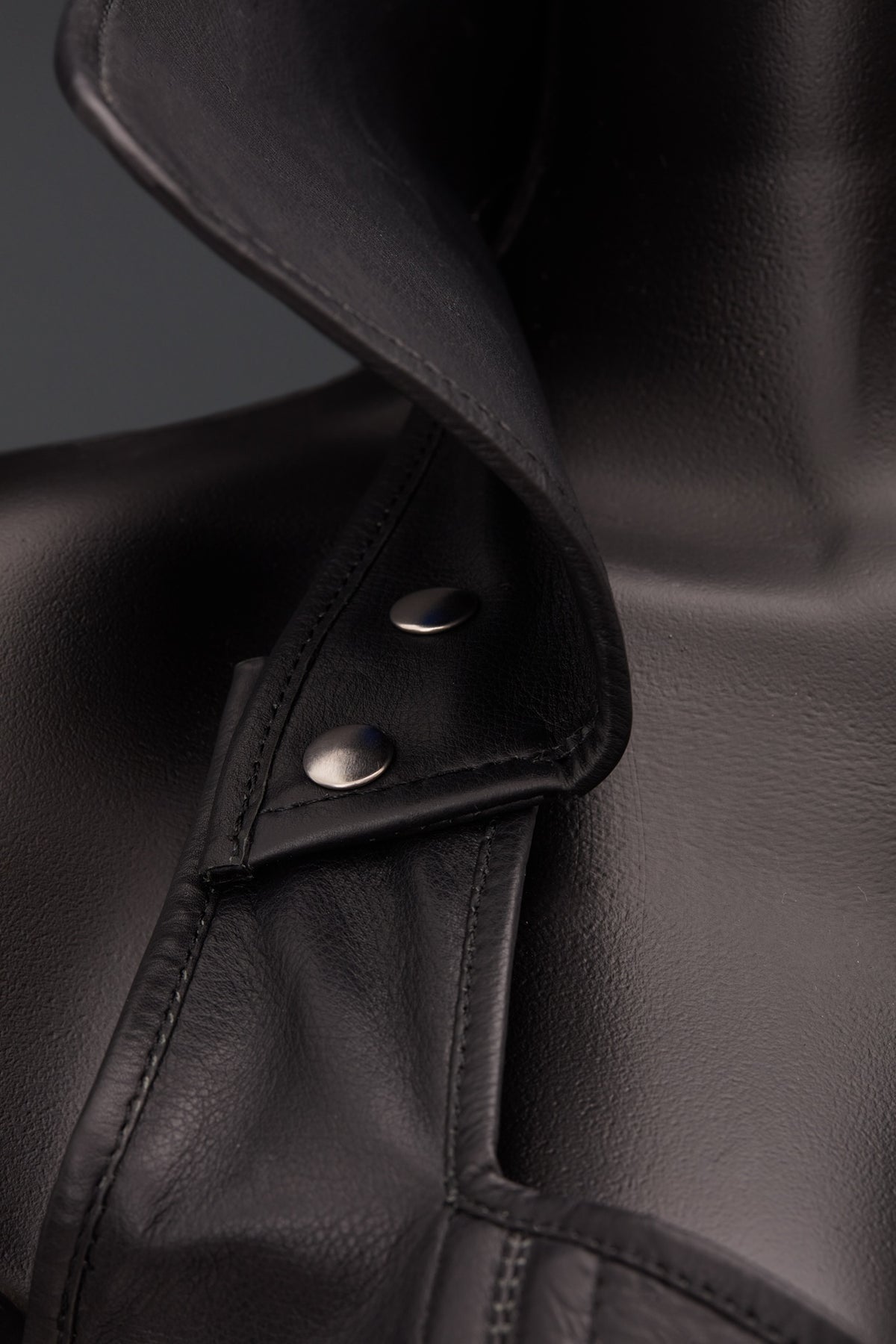Close-up of a black leather corset with a pointed collar and silver snap button detail, shown on a dark mannequin against a dark gray background.
