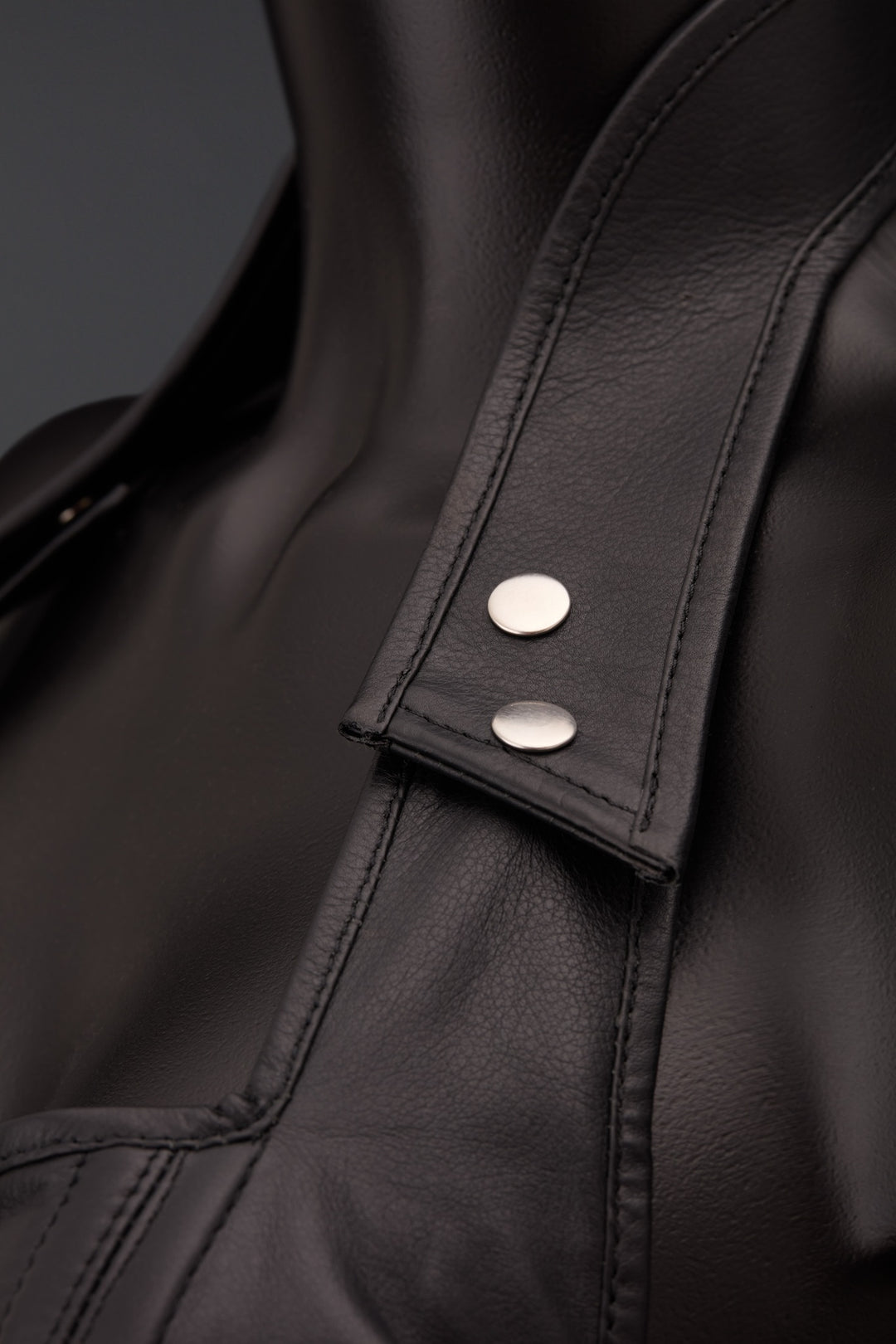 Close-up of a black leather corset featuring a pointed collar with two silver-tone snap buttons on a tab, set against a dark gray background.
