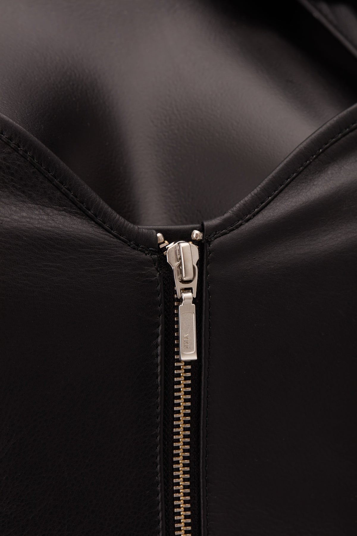 Close-up of a black leather zipper detail with a silver-toned pull tab on a black background.