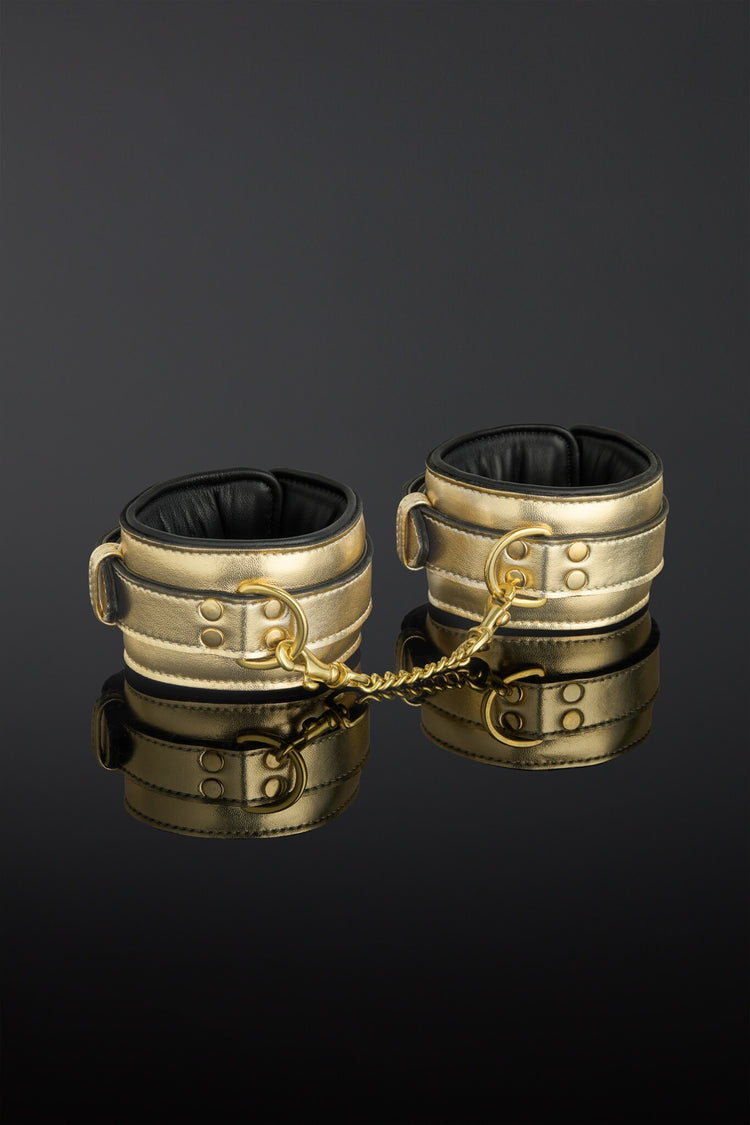 A pair of metallic gold double-strap leather cuffs with black interior lining and a gold chain link, displayed on a reflective black surface.