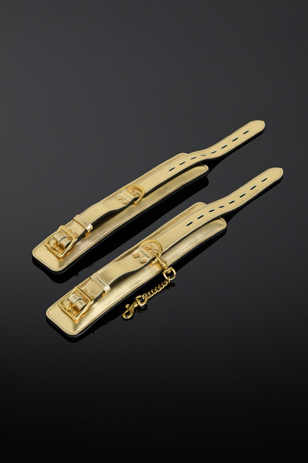 Two gold-tone leather cuff strap adjusters with metal buckles and a short chain, displayed on a reflective black background.