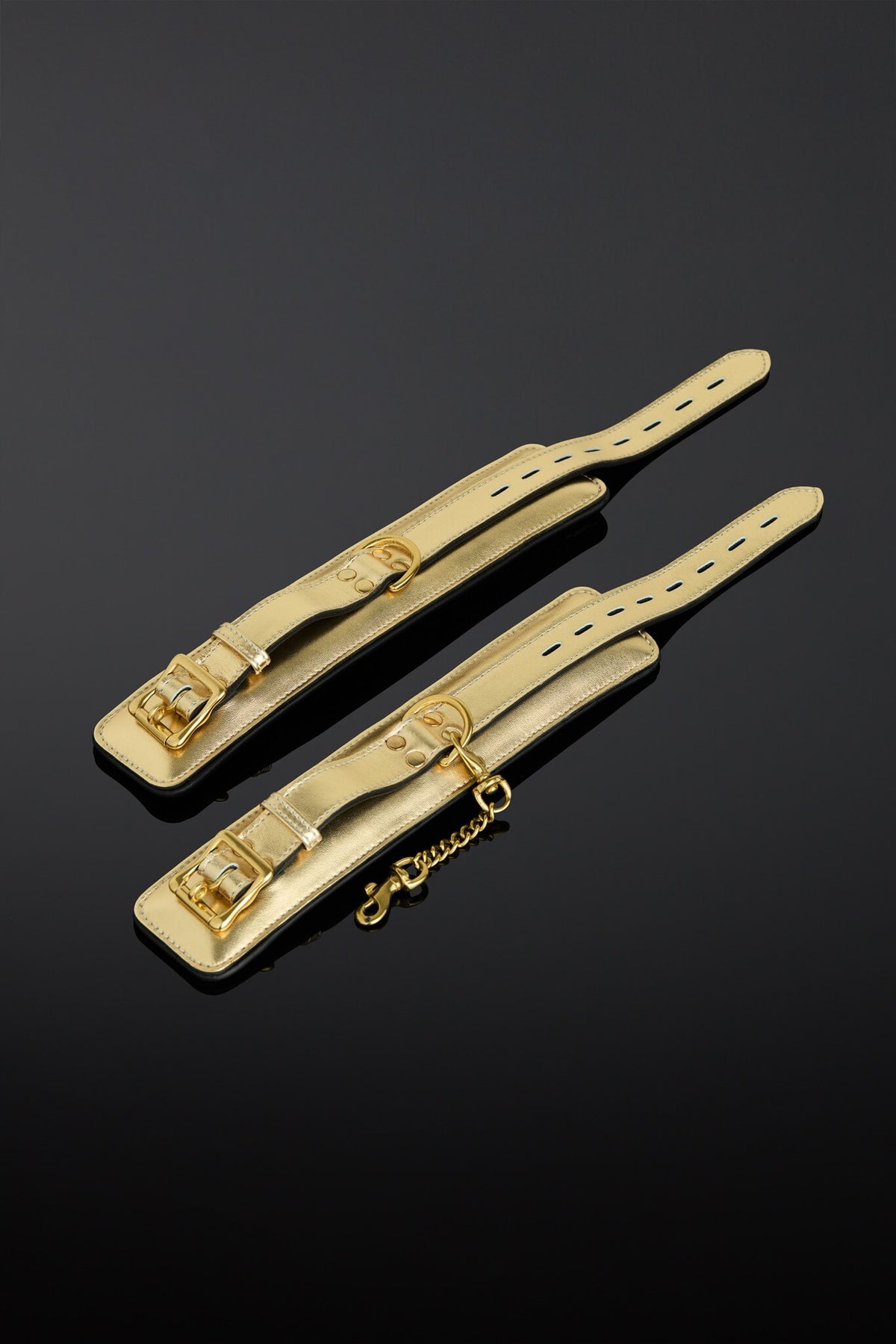 Two gold-tone leather cuff strap adjusters with metal buckles and a short chain, displayed on a reflective black background.