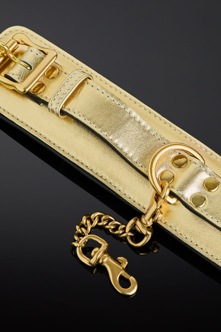 A close-up of a metallic gold leather cuffs with matching gold hardware, including a buckle, rivets, and a hanging chain clasp, set against a dark reflective background.