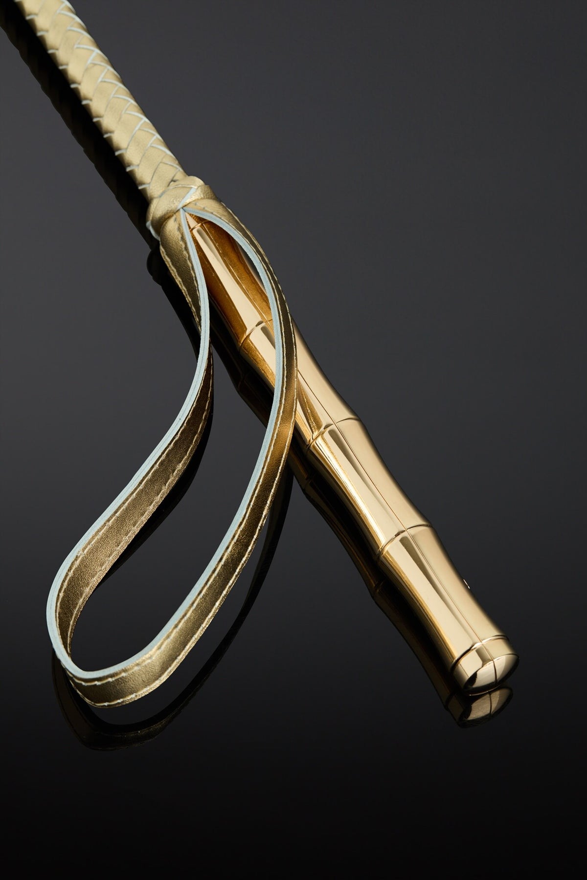 A close-up studio shot of a gold-toned leather riding crop or whip with a smooth cylindrical handle, resting on a glossy black reflective surface.