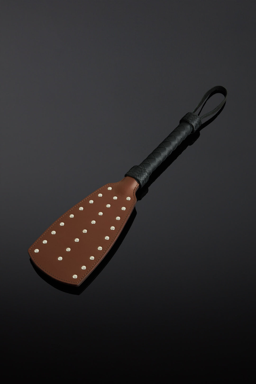 Photo of a brown leather paddle with black leather braided handle. There are multiple rivets on the face of the paddle. 