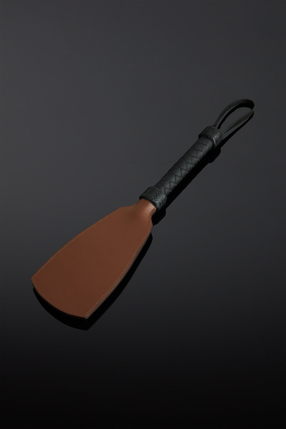 Photo of a brown leather paddle with black leather braided handle.  On a black background