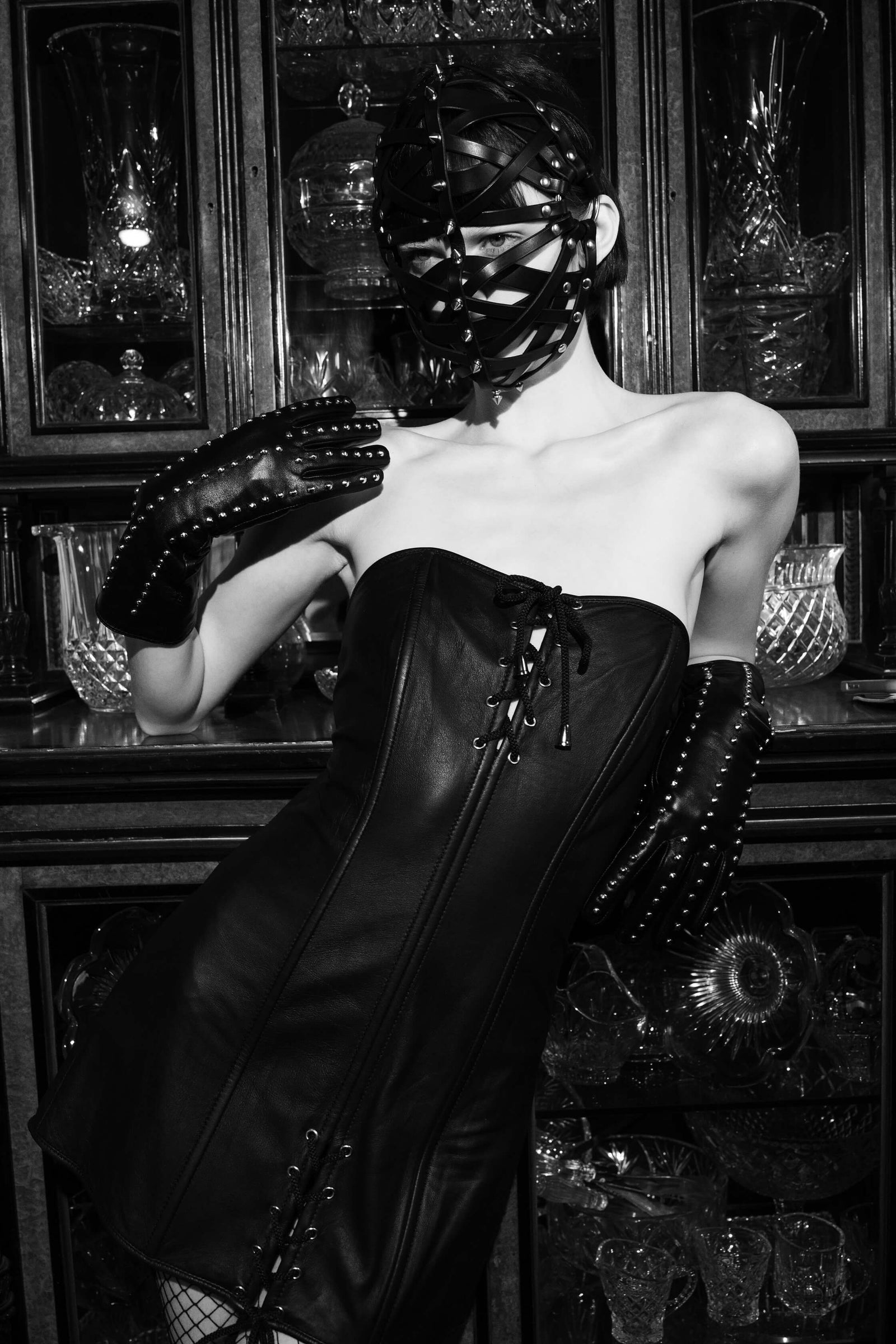 Photo of House of SXN model wearing Copulatta mask.  She is standing in front of an antique glassware wooden cabinet. She is dressed in black leather gloves and black leather dress. 