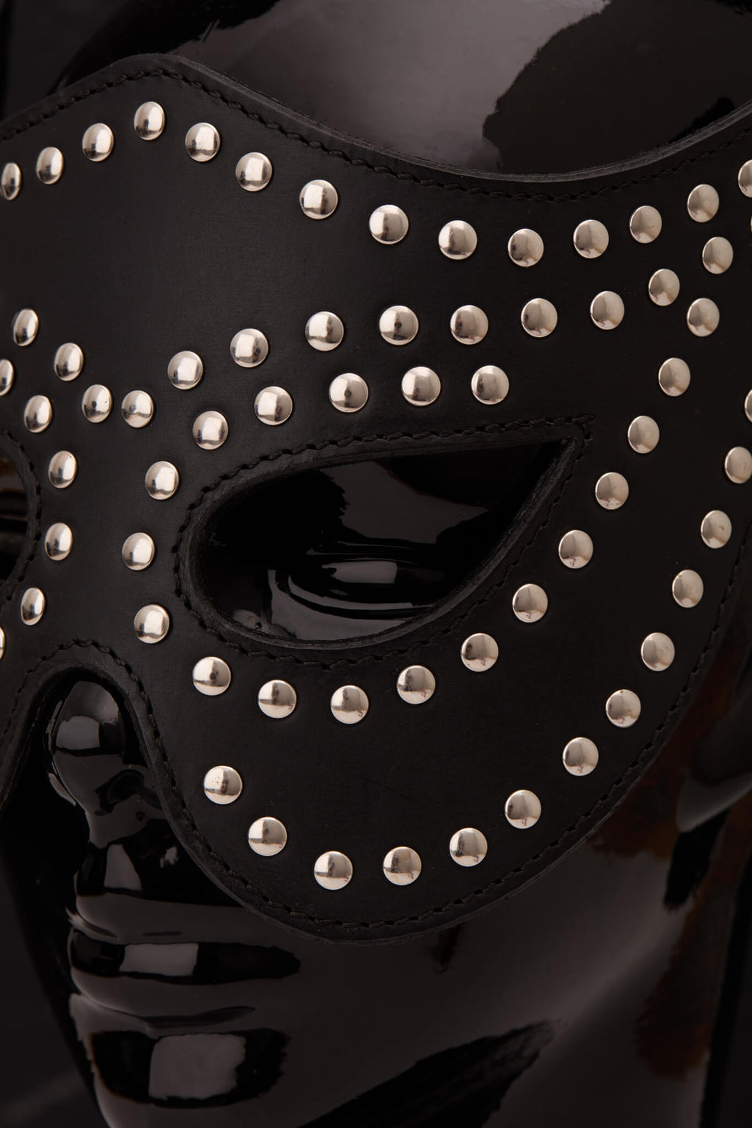 A black leather masquerade mask adorned with silver studs, displayed on a glossy black mannequin head against a dark gray background.