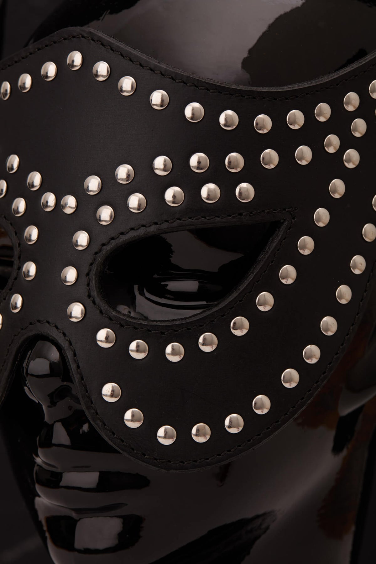 A black leather masquerade mask adorned with silver studs, displayed on a glossy black mannequin head against a dark gray background.