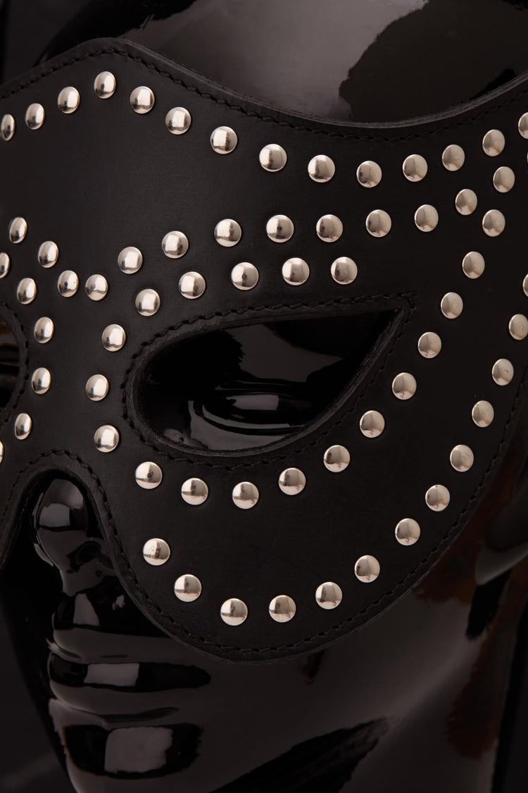 A black leather masquerade mask adorned with silver studs, displayed on a glossy black mannequin head against a dark gray background.