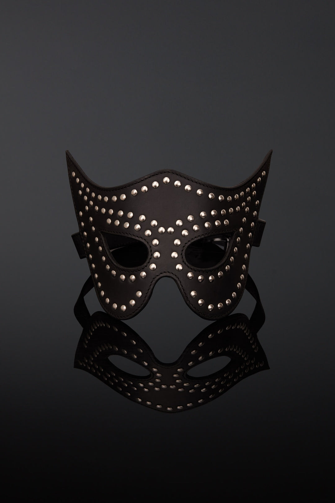A black leather masquerade mask adorned with silver studs, displayed on a reflective black surface against a dark gray background.
