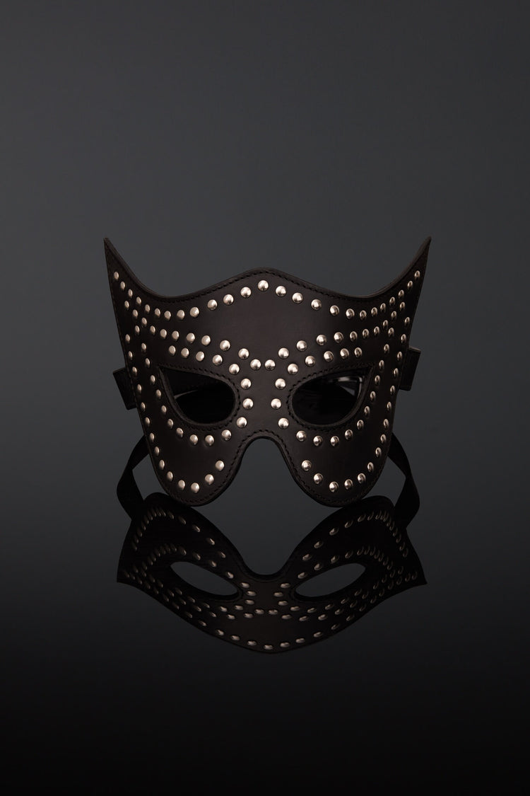 A black leather masquerade mask adorned with silver studs, displayed on a reflective black surface against a dark gray background.