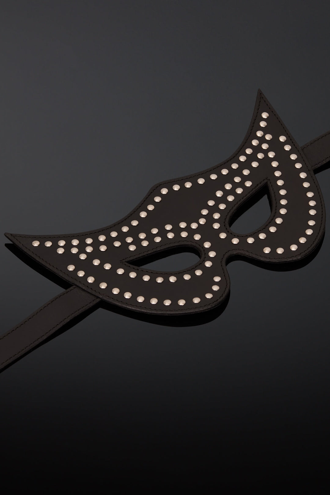 A black leather masquerade mask adorned with silver studs, displayed on a glossy black mannequin head against a dark gray background.