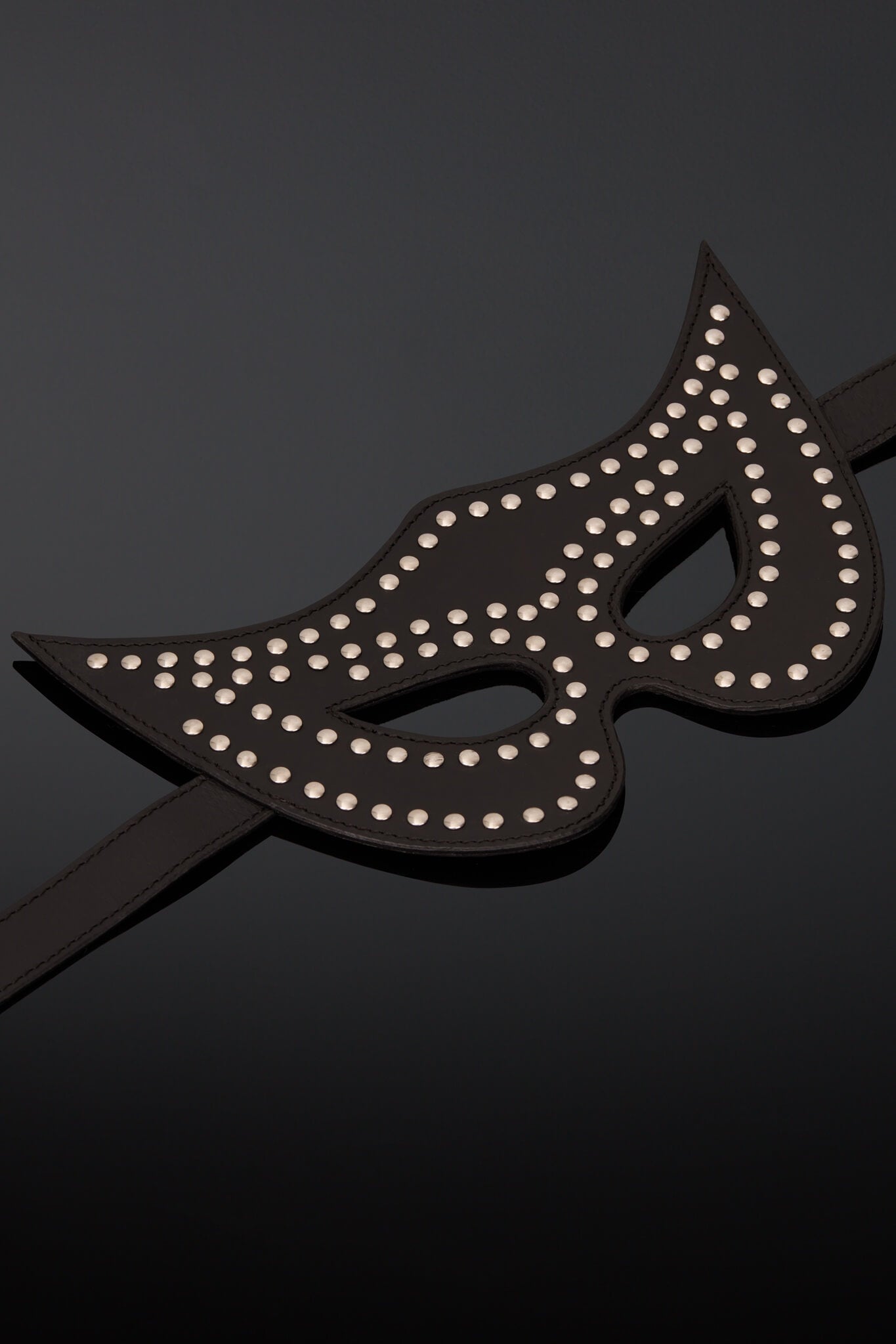 A black leather masquerade mask adorned with silver studs, displayed on a glossy black mannequin head against a dark gray background.