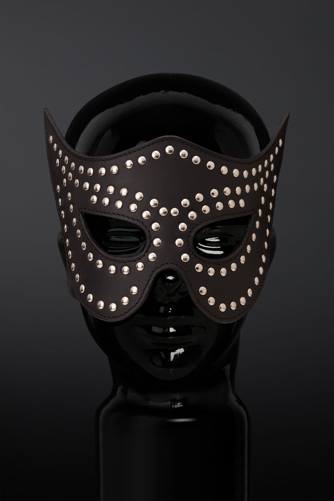 A black leather masquerade mask adorned with silver studs, displayed on a glossy black mannequin head against a dark gray background.