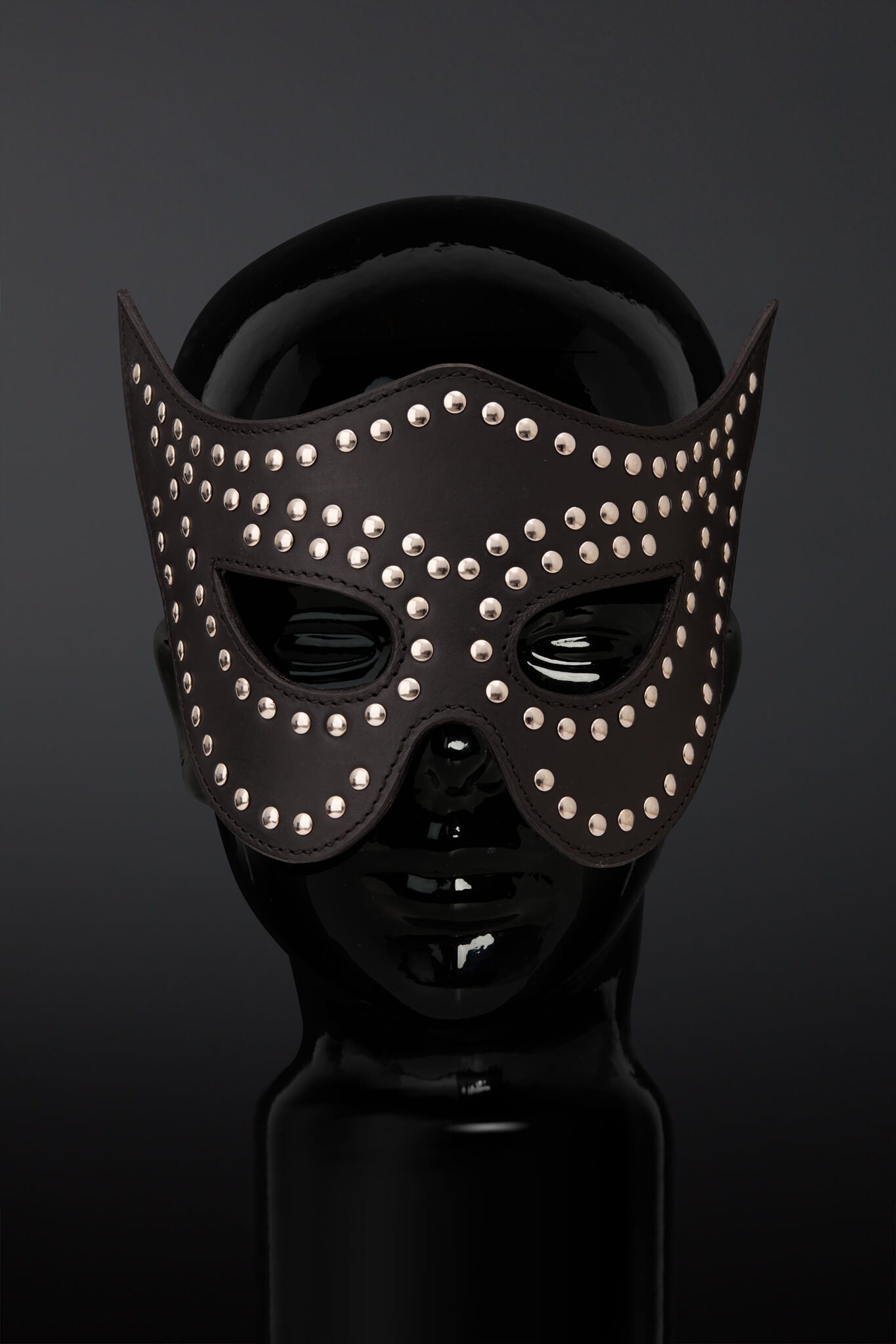 A black leather masquerade mask adorned with silver studs, displayed on a glossy black mannequin head against a dark gray background.