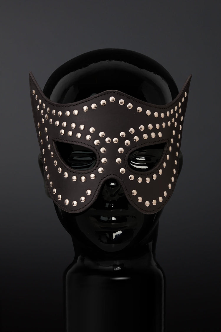 A black leather masquerade mask adorned with silver studs, displayed on a glossy black mannequin head against a dark gray background.