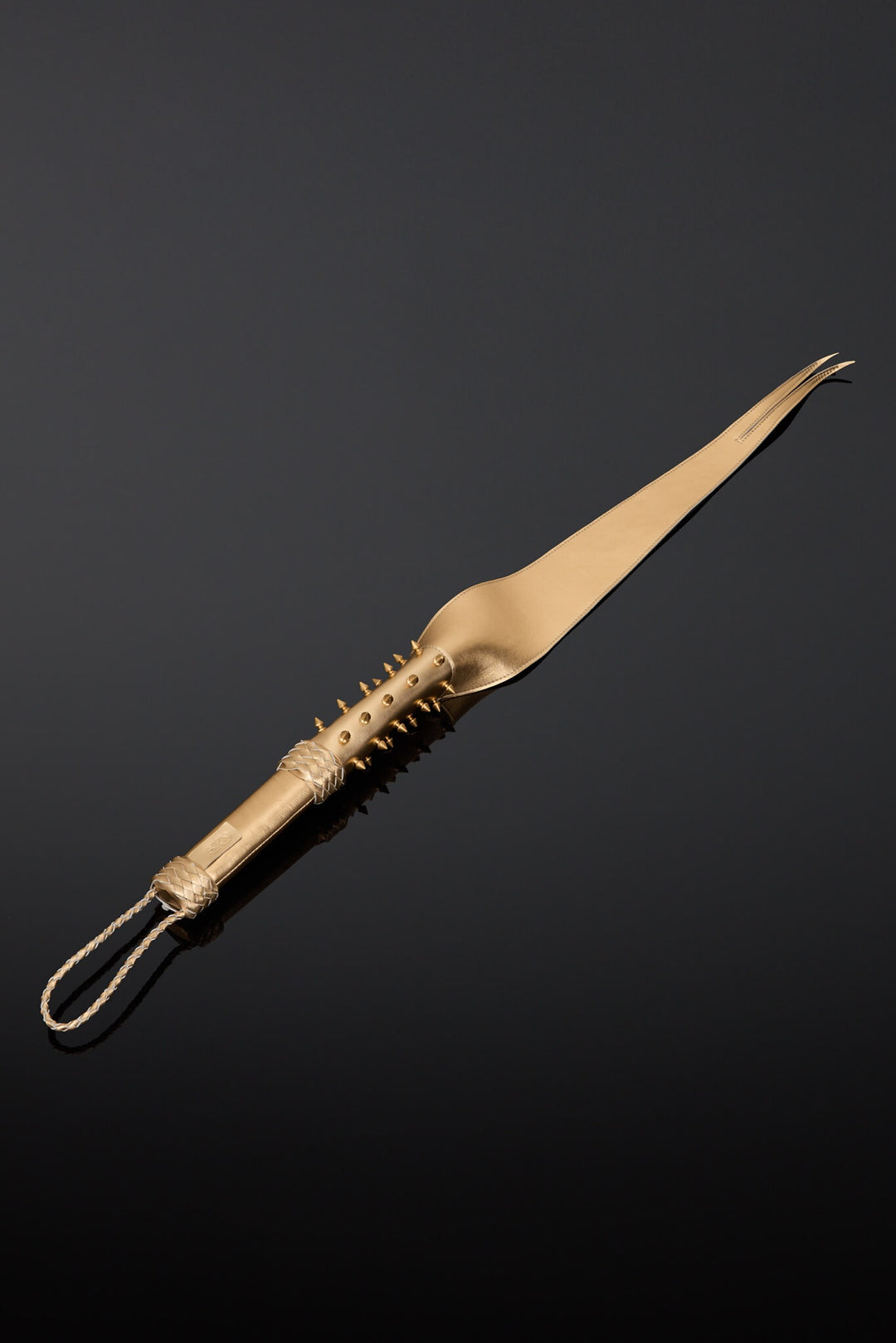 A gold-colored leather dragon tail whip with a textured, spiked handle and a hanging loop, displayed diagonally on a reflective black surface.