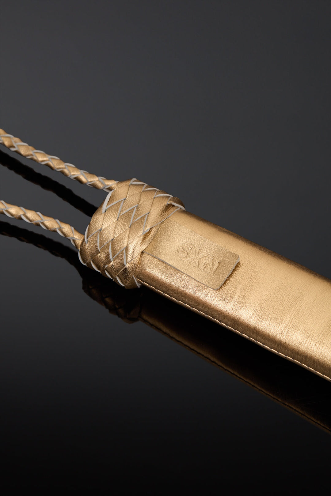 A close-up of a gold-toned leather accessory with braided rope details and a small embossed brand patch, photographed on a dark reflective surface.