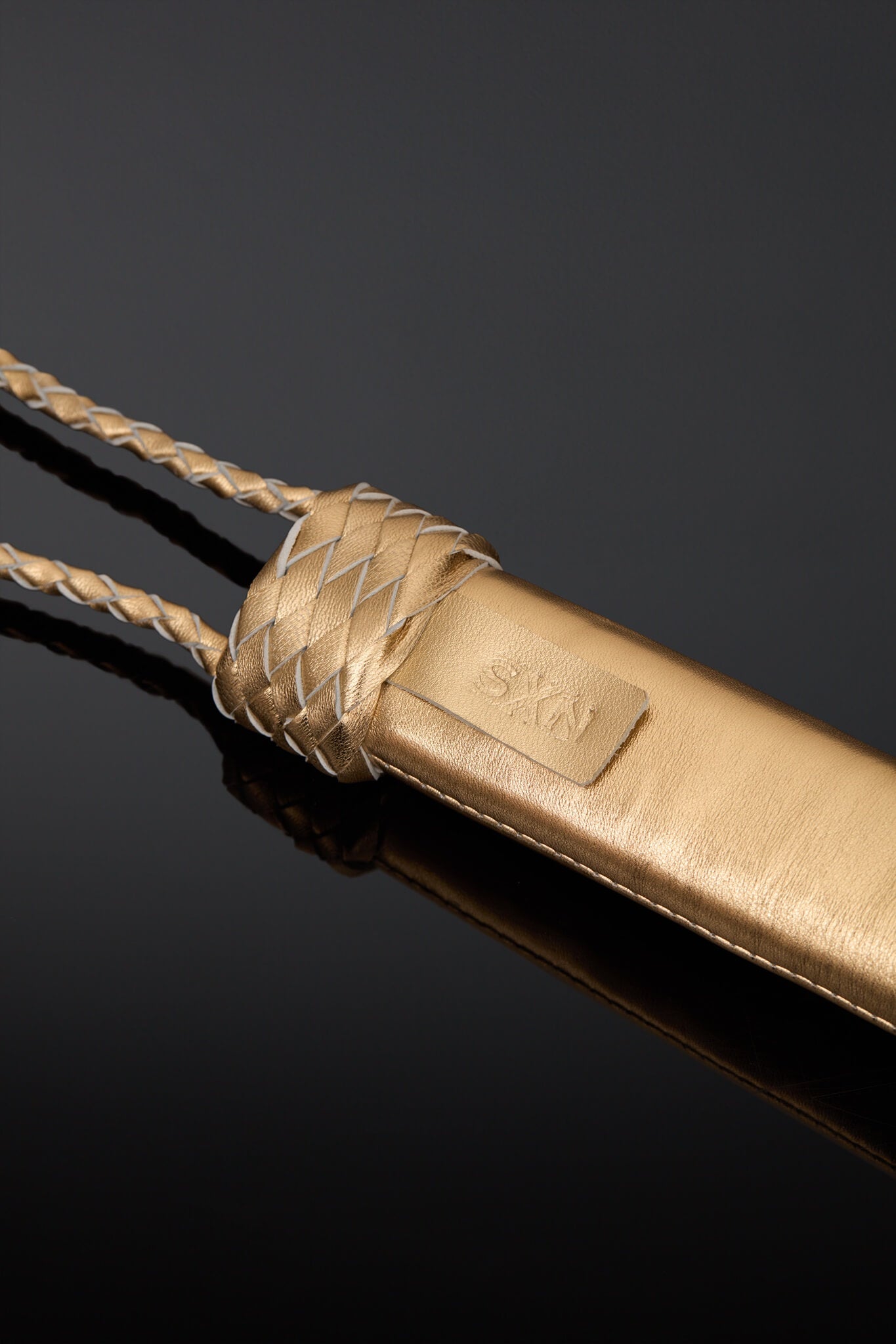 A close-up of a gold-toned leather accessory with braided rope details and a small embossed brand patch, photographed on a dark reflective surface.
