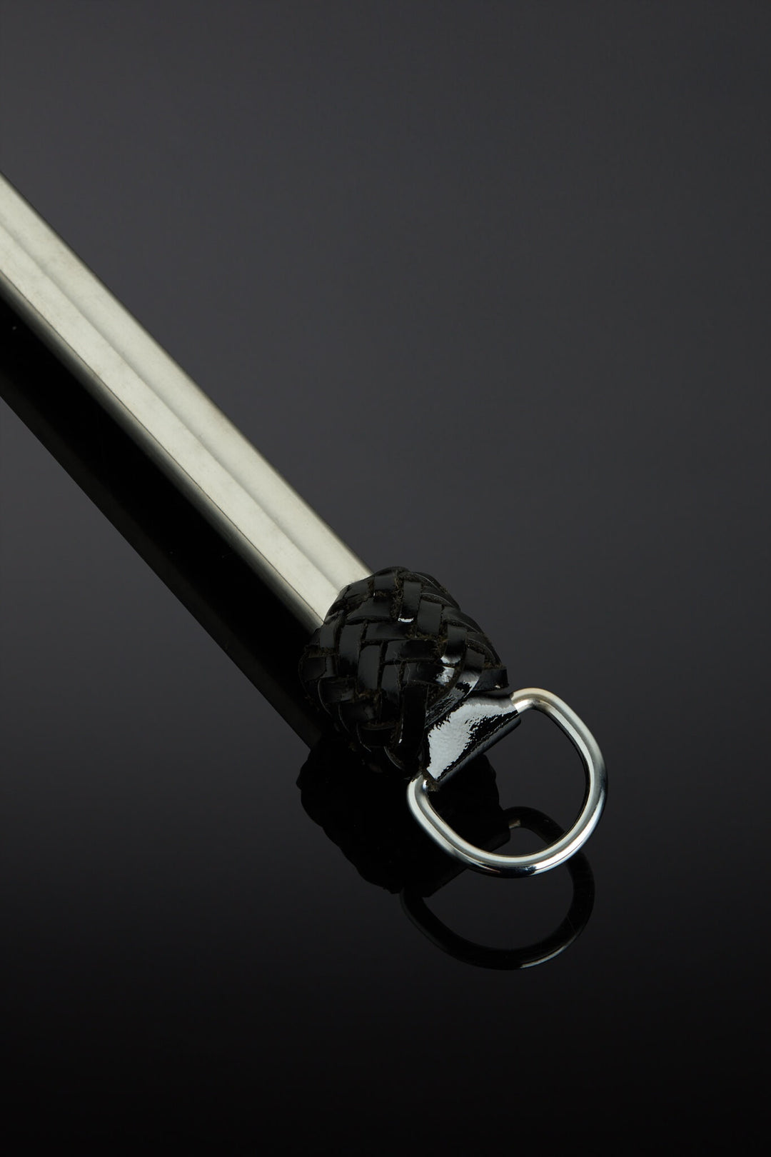 A close-up of a black leather-wrapped martial arts stick with a silver metal D-ring, set against a dark reflective background.