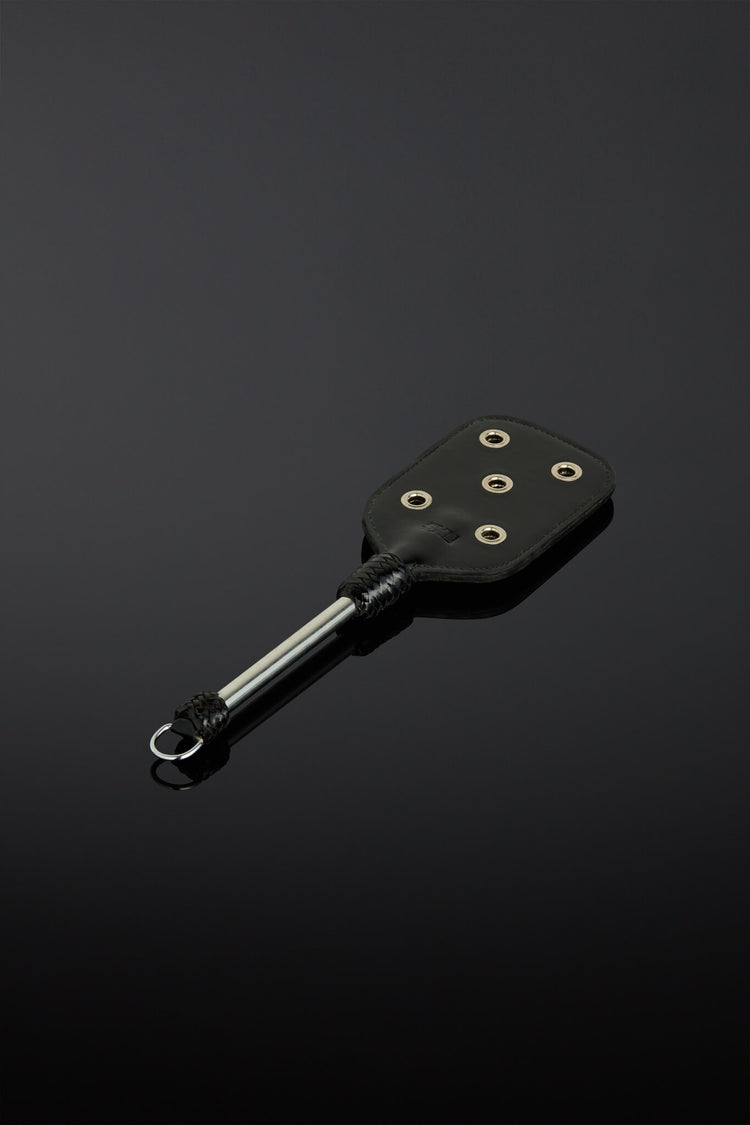 A black leather paddle-style accessory with silver metal grommets and a silver metal rod with a black knob, photographed on a dark reflective surface.