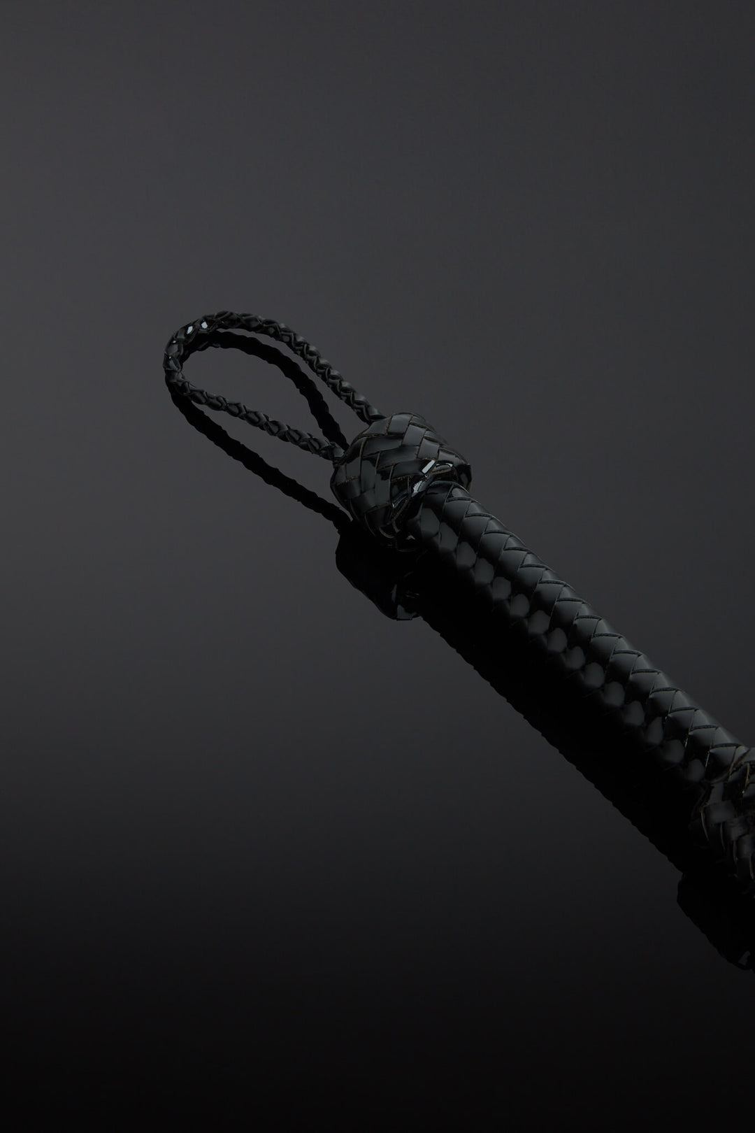 Close-up of a black patent leather flogger with a knot on the handle on a dark background