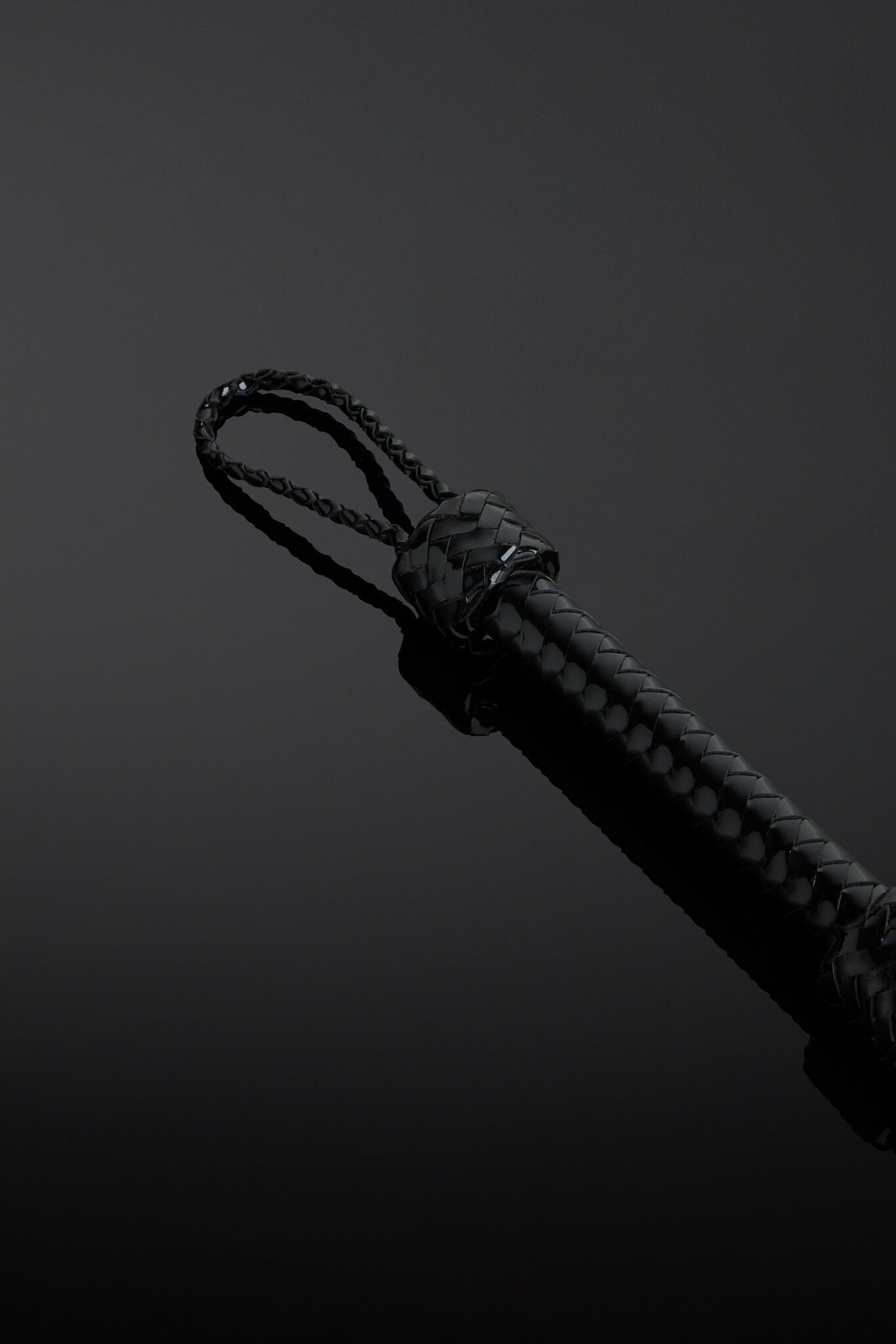 Close-up of a black patent leather flogger with a knot on the handle on a dark background