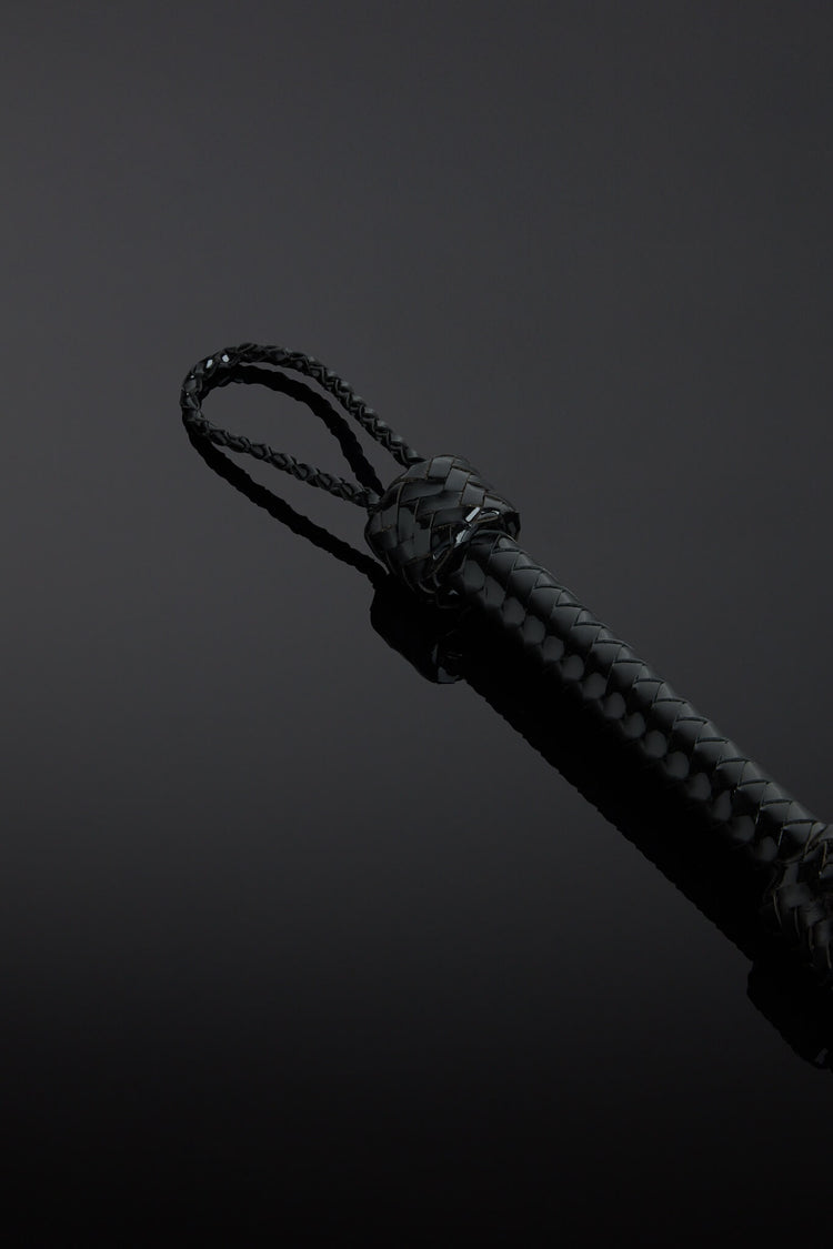 Close-up of a black patent leather flogger with a knot on the handle on a dark background